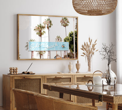 Colorful travel photography capturing the iconic 'Solana Beach' sign, a quintessential piece of Surf Signs San Diego art. The retro light blue sign, adorned with white cursive script and a unique steer head logo, is set against a backdrop of towering palm trees and a bright, hazy California sky. This Southern California surf beach sign evokes strong summer vibes and is perfect for coastal decor. Photograph in minimalist framed mock up.