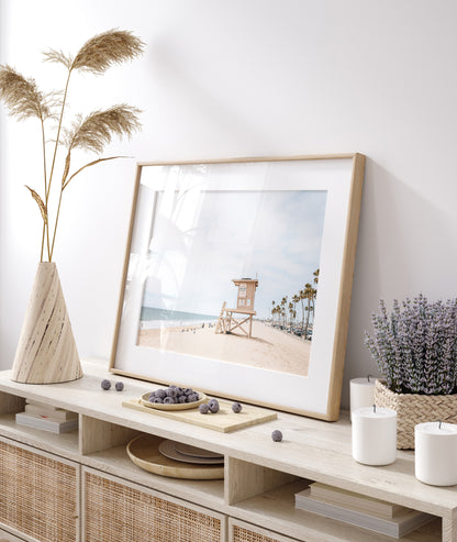 A wide landscape coastal photography view of Newport Beach in Southern California, featuring a row of tall palm trees, a lifeguard tower, and people enjoying the calm beach. Photograph in minimalist mock up.