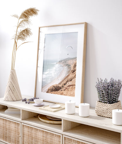 Fine art of Torrey Pines, San Diego California vertical print of hazy high-angle landscape beach photography captures a light blue ocean meeting a sandy beach with many beach goers. To the right, tall tan and brown cliffs rise as iconic coastal scenery. A white paraglider with a pilot flies in the upper right against a pale pastel sky, a travel photography moment. The scene emphasizes the cliffs, beach, and the paraglider's presence in the expansive large beach art view. Photograph in minimalist mock up
