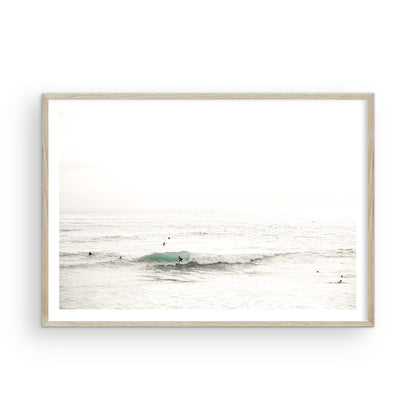 Fine art of horizontal landscape print by Southern California photographer Kim Dybczak of Salt Creek Prints, featuring a bright image captures a pale sandy beach giving way to a vast, light gray ocean with gentle waves. Several surfers are scattered across the water, some sitting on their boards and others riding small waves. The sky is a uniform, pale white, creating a soft, overcast atmosphere, located in Windansea Beach in La Jolla, California. Photograph in framed minimalist mock up
