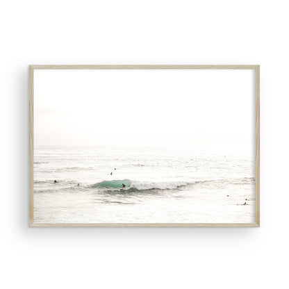Fine art of horizontal landscape print by Southern California photographer Kim Dybczak of Salt Creek Prints, featuring a bright image captures a pale sandy beach giving way to a vast, light gray ocean with gentle waves. Several surfers are scattered across the water, some sitting on their boards and others riding small waves. The sky is a uniform, pale white, creating a soft, overcast atmosphere, located in Windansea Beach in La Jolla, California. Photograph in framed minimalist mock up