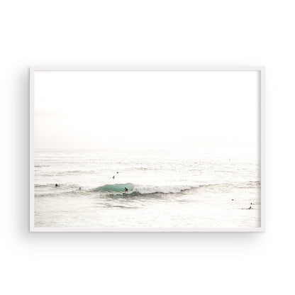 Fine art of horizontal landscape print by Southern California photographer Kim Dybczak of Salt Creek Prints, featuring a bright image captures a pale sandy beach giving way to a vast, light gray ocean with gentle waves. Several surfers are scattered across the water, some sitting on their boards and others riding small waves. The sky is a uniform, pale white, creating a soft, overcast atmosphere, located in Windansea Beach in La Jolla, California. Photograph in framed minimalist mock up