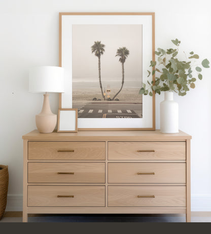 Vertical coastal photography of two leaning palm trees on a hazy Oceanside beach, with ocean waves in the background and a crosswalk in the foreground. Photograph in minimalist mock up.