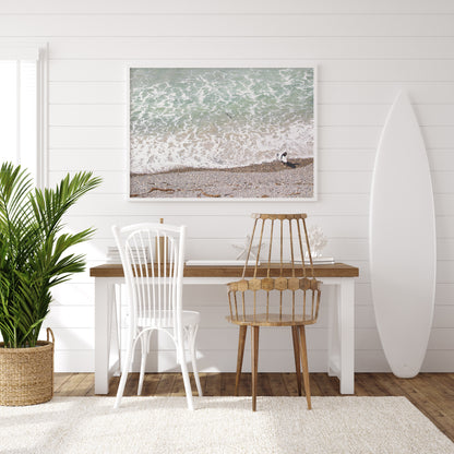 Horizontal surf print of a surfer with a surfboard walking along a rocky beach next to foamy ocean waves. This surfing art captures a unique coastline. Photograph in minimalist mock up.