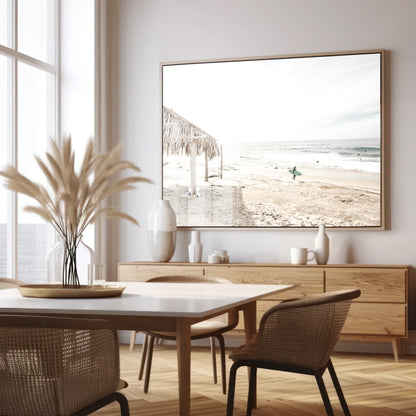 Fine art of WindanSea Beach, La Jolla California horizontal landscape print by Southern California photographer Kim Dybczak of Salt Creek Prints, featuring a bright, slightly desaturated image shows Windansea Beach in La Jolla, California. A thatched beach hut stands on the sandy shore in the left foreground. Further down the beach, a surfer carrying a teal surfboard walks towards the ocean where several other surfers are visible in the water. Photograph in minimalist framed dining room mock up