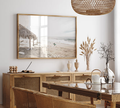 Fine art of WindanSea Beach, La Jolla California horizontal landscape print by Southern California photographer Kim Dybczak of Salt Creek Prints, featuring a bright, slightly desaturated image shows Windansea Beach in La Jolla, California. A thatched beach hut stands on the sandy shore in the left foreground. Further down the beach, a surfer carrying a teal surfboard walks towards the ocean where several other surfers are visible in the water. Photograph in minimalist framed dining mock up