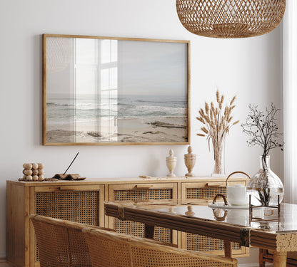 Fine art of horizontal landscape print by Southern California photographer Kim Dybczak of Salt Creek Prints, featuring a bright image captures a pale sandy beach giving way to a vast, light gray ocean with gentle waves. Several surfers are scattered across the water, some sitting on their boards and others riding small waves. The sky is a uniform, pale white, creating a soft, overcast atmosphere, located in Windansea Beach in La Jolla, California. Photograph in framed minimalist dining mock up
