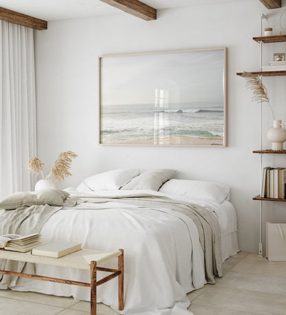 Fine art of horizontal landscape print by Southern California photographer Kim Dybczak of Salt Creek Prints, featuring a bright image captures a pale sandy beach giving way to a vast, light gray ocean with gentle waves. Several surfers are scattered across the water, some sitting on their boards and others riding small waves. The sky is a uniform, pale white, creating a soft, overcast atmosphere, located in Windansea Beach in La Jolla, California. Photograph in framed minimalist bedroom mock up