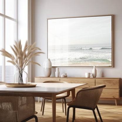 Fine art of horizontal landscape print by Southern California photographer Kim Dybczak of Salt Creek Prints, featuring a bright image captures a pale sandy beach giving way to a vast, light gray ocean with gentle waves. Several surfers are scattered across the water, some sitting on their boards and others riding small waves. The sky is a uniform, pale white, creating a soft, overcast atmosphere, located in Windansea Beach in La Jolla, California. Photograph in framed minimalist dining room mock up