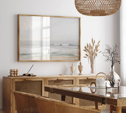 Fine art of horizontal landscape print by Southern California photographer Kim Dybczak of Salt Creek Prints, featuring a bright image captures a pale sandy beach giving way to a vast, light gray ocean with gentle waves. Several surfers are scattered across the water, some sitting on their boards and others riding small waves. The sky is a uniform, pale white, creating a soft, overcast atmosphere, located in Windansea Beach in La Jolla, California. Photograph in framed minimalist mock up