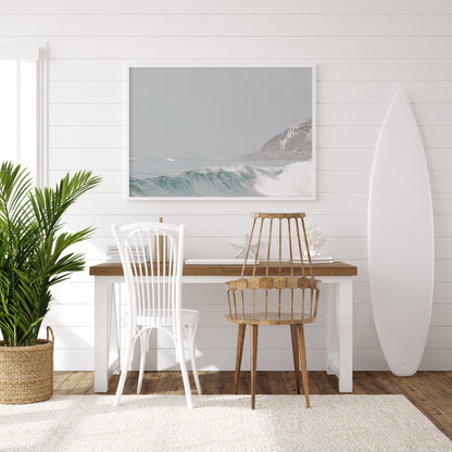 Neutral ocean print featuring a beautifully captured turquoise wave cresting with white seafoam, embodying minimalist surf art. This horizontal coastal photography showcases soft, pastel tones under a hazy sky. In the background, a rugged cliffside staircase on the right meets the sea, with faint distant surfers in the water, suggesting the expansive Southern California coastline. A tranquil piece of surf wall art, perfect for a coastal theme. Photograph in surfers room minimalist mock up.