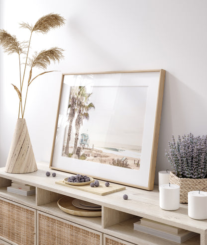 Dreamy coastal palm tree print featuring two tall palm trees with shaggy trunks in the foreground and a pastel blue lifeguard tower in the distance. This horizontal tropical print is captured at Seaside Beach on the Southern California coast, is bathed in bright, hazy sunlight, creating a pastel beach art effect. Evokes serene summer vibes, perfect for beach house decor. Photograph in minimalist mock up.