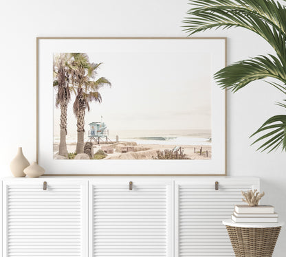 Dreamy coastal palm tree print featuring two tall palm trees with shaggy trunks in the foreground and a pastel blue lifeguard tower in the distance. This horizontal tropical print is captured at Seaside Beach on the Southern California coast, is bathed in bright, hazy sunlight, creating a pastel beach art effect. Evokes serene summer vibes, perfect for beach house decor. Photograph in minimalist mock up.