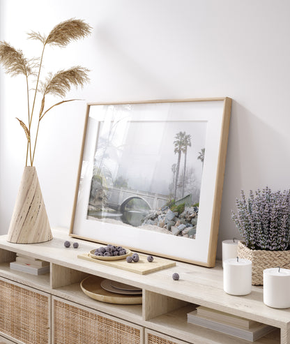 Landscape coastal photography of a concrete arch bridge disappearing into a dense fog, flanked by palm trees and rocky shores. This calm beach print evokes a mysterious seascape. Photograph in minimalist mock up.