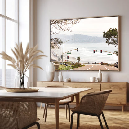 A hazy eye-level shot of San Diego Streets looks down a sloping road featuring red traffic lights and "Carmel Valley" signs, capturing a Local Surf Print vibe. The foreground of this California Wall Art showcases landscape and cars, framed by tree branches at the top, adding to the San Diego Photo aesthetic. The lighting suggests a hazy daytime, perfect for Minimalist Beach Wall Art. This horizontal San Diego print makes a great piece of horizontal beach wall decor. Photograph in minimalist dining mock up