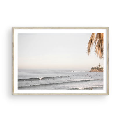 Landscape beach photography of Tourmaline Beach, Pacific Beach, California, with waves and many surfers, framed by a palm tree in the corner. This ocean print captures casual surf vibes. Photograph in minimalist mock up.