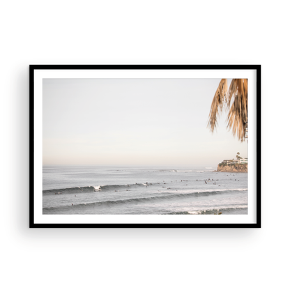 Landscape beach photography of Tourmaline Beach, Pacific Beach, California, with waves and many surfers, framed by a palm tree in the corner. This ocean print captures casual surf vibes. Photograph in minimalist mock up.