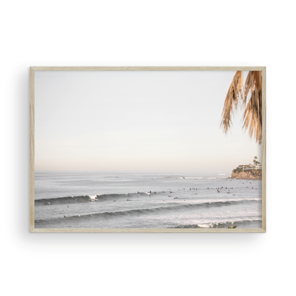Landscape beach photography of Tourmaline Beach, Pacific Beach, California, with waves and many surfers, framed by a palm tree in the corner. This ocean print captures casual surf vibes. Photograph in minimalist mock up.