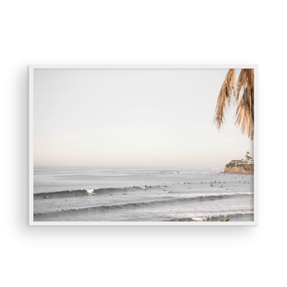 Landscape beach photography of Tourmaline Beach, Pacific Beach, California, with waves and many surfers, framed by a palm tree in the corner. This ocean print captures casual surf vibes. Photograph in minimalist mock up.