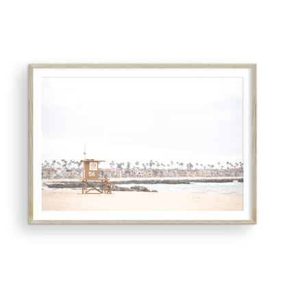 Landscape beach photography of Newport Beach, featuring lifeguard tower 56 on the sand with ocean waves and coastal houses in the background. A great Newport Beach photography print. Photograph in minimalist mock up