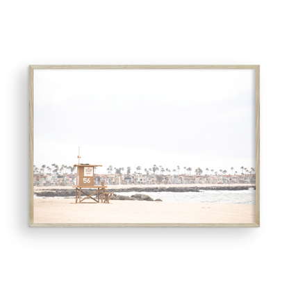 Landscape beach photography of Newport Beach, featuring lifeguard tower 56 on the sand with ocean waves and coastal houses in the background. A great Newport Beach photography print. Photograph in minimalist mock up