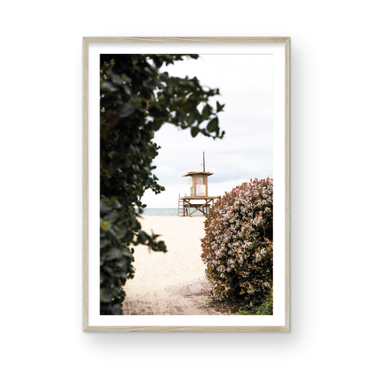 Vertical coastal photography of a Newport Beach lifeguard tower on the sand, framed by lush green and flowering foliage. Photograph in minimalist mock up.