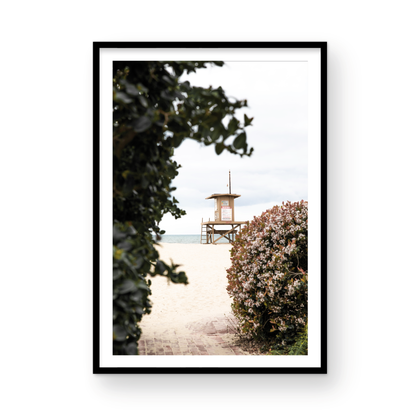 Vertical coastal photography of a Newport Beach lifeguard tower on the sand, framed by lush green and flowering foliage. Photograph in minimalist mock up.