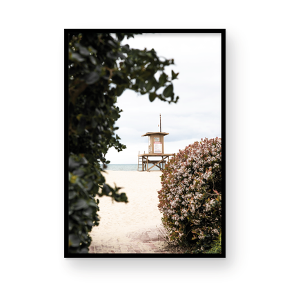 Vertical coastal photography of a Newport Beach lifeguard tower on the sand, framed by lush green and flowering foliage. Photograph in minimalist mock up.