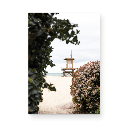 Vertical coastal photography of a Newport Beach lifeguard tower on the sand, framed by lush green and flowering foliage. Photograph in minimalist mock up.