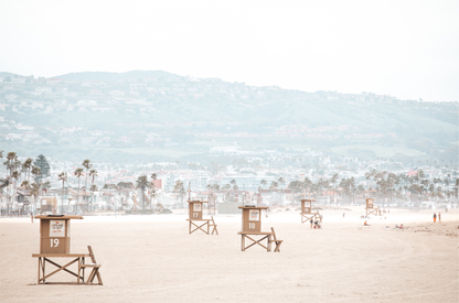 Landscape beach photography of Santa Monica State Beach with multiple lifeguard towers on the vast sandy expanse and hazy hills in the background. 