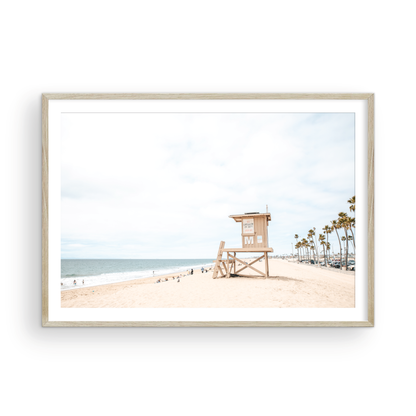 A wide landscape coastal photography view of Newport Beach in Southern California, featuring a row of tall palm trees, a lifeguard tower, and people enjoying the calm beach. Photograph in minimalist mock up.