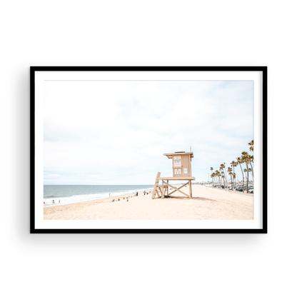 A wide landscape coastal photography view of Newport Beach in Southern California, featuring a row of tall palm trees, a lifeguard tower, and people enjoying the calm beach. Photograph in minimalist mock up.