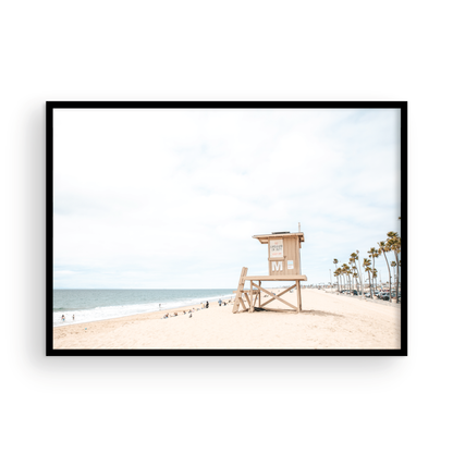 A wide landscape coastal photography view of Newport Beach in Southern California, featuring a row of tall palm trees, a lifeguard tower, and people enjoying the calm beach. Photograph in minimalist mock up.