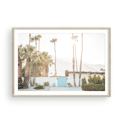 Coastal print of a Palm Springs home with a bright blue door, surrounded by tall palm trees and distant mountains, embodying summer vibes wall art. Photograph in minimalist mock up.