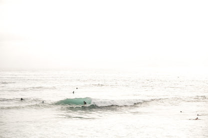 Fine art of horizontal landscape print by Southern California photographer Kim Dybczak of Salt Creek Prints, featuring a bright image captures a pale sandy beach giving way to a vast, light gray ocean with gentle waves. Several surfers are scattered across the water, some sitting on their boards and others riding small waves. The sky is a uniform, pale white, creating a soft, overcast atmosphere, located in Windansea Beach in La Jolla, California. 