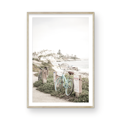Fine art of horizontal landscape print by Southern California photographer Kim Dybczak of Salt Creek Prints, the image shows a light blue pastel beach cruiser leaning against a wooden post and chain fence. Another bicycle is visible behind it. The beach is in the background with people and a beach hut, located in Windansea Beach in La Jolla, California. Photograph in framed minimalist mock up