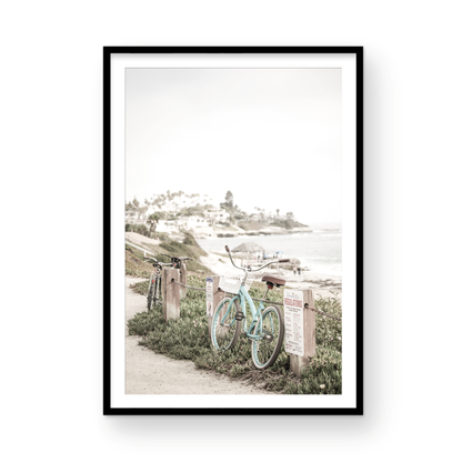 Fine art of horizontal landscape print by Southern California photographer Kim Dybczak of Salt Creek Prints, the image shows a light blue pastel beach cruiser leaning against a wooden post and chain fence. Another bicycle is visible behind it. The beach is in the background with people and a beach hut, located in Windansea Beach in La Jolla, California. Photograph in framed minimalist mock up
