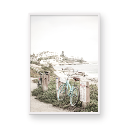 Fine art of horizontal landscape print by Southern California photographer Kim Dybczak of Salt Creek Prints, the image shows a light blue pastel beach cruiser leaning against a wooden post and chain fence. Another bicycle is visible behind it. The beach is in the background with people and a beach hut, located in Windansea Beach in La Jolla, California. Photograph in framed minimalist mock up