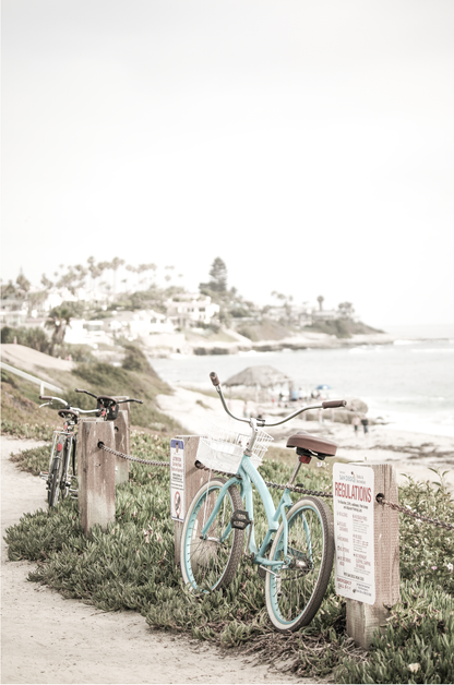 Fine art of horizontal landscape print by Southern California photographer Kim Dybczak of Salt Creek Prints, the image shows a light blue pastel beach cruiser leaning against a wooden post and chain fence. Another bicycle is visible behind it. The beach is in the background with people and a beach hut, located in Windansea Beach in La Jolla, California. Photograph in framed minimalist mock up