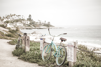 Fine art of horizontal landscape print by Southern California photographer Kim Dybczak of Salt Creek Prints, the image shows a light blue pastel beach cruiser leaning against a wooden post and chain fence. Another bicycle is visible behind it. The beach is in the background with people and a beach hut, located in Windansea Beach in La Jolla, California.