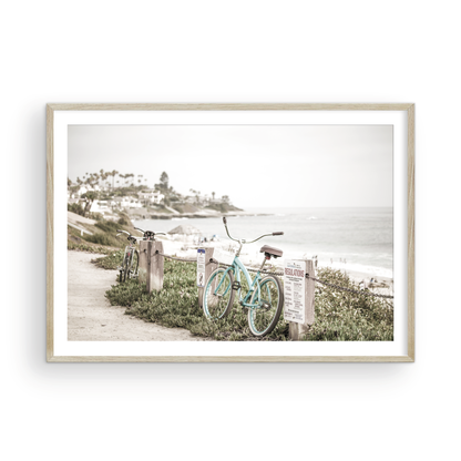 Fine art of horizontal landscape print by Southern California photographer Kim Dybczak of Salt Creek Prints, the image shows a light blue pastel beach cruiser leaning against a wooden post and chain fence. Another bicycle is visible behind it. The beach is in the background with people and a beach hut, located in Windansea Beach in La Jolla, California. Photograph in framed minimalist mock up