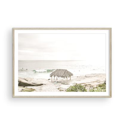 Fine art of WindanSea Beach, La Jolla California horizontal landscape print by Southern California photographer Kim Dybczak of Salt Creek Prints, featuring a bright, slightly desaturated image shows Windansea Beach in La Jolla, California. A thatched beach hut stands on the sandy shore in the left foreground. Further down the beach, a surfer carrying a teal surfboard walks towards the ocean where several other surfers are visible in the water. Photograph in minimalist framed mock up