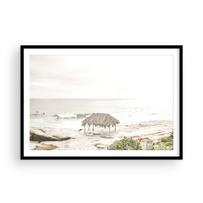 Fine art of WindanSea Beach, La Jolla California horizontal landscape print by Southern California photographer Kim Dybczak of Salt Creek Prints, featuring a bright, slightly desaturated image shows Windansea Beach in La Jolla, California. A thatched beach hut stands on the sandy shore in the left foreground. Further down the beach, a surfer carrying a teal surfboard walks towards the ocean where several other surfers are visible in the water. Photograph in minimalist framed mock up