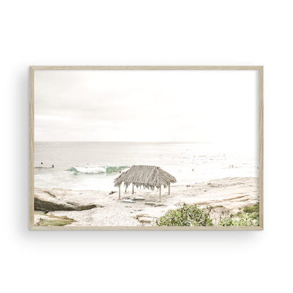 Fine art of WindanSea Beach, La Jolla California horizontal landscape print by Southern California photographer Kim Dybczak of Salt Creek Prints, featuring a bright, slightly desaturated image shows Windansea Beach in La Jolla, California. A thatched beach hut stands on the sandy shore in the left foreground. Further down the beach, a surfer carrying a teal surfboard walks towards the ocean where several other surfers are visible in the water. Photograph in minimalist framed mock up