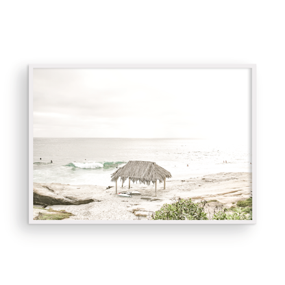 Fine art of WindanSea Beach, La Jolla California horizontal landscape print by Southern California photographer Kim Dybczak of Salt Creek Prints, featuring a bright, slightly desaturated image shows Windansea Beach in La Jolla, California. A thatched beach hut stands on the sandy shore in the left foreground. Further down the beach, a surfer carrying a teal surfboard walks towards the ocean where several other surfers are visible in the water. Photograph in minimalist framed mock up