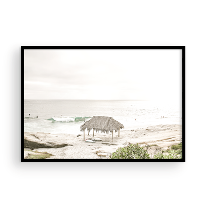 Fine art of WindanSea Beach, La Jolla California horizontal landscape print by Southern California photographer Kim Dybczak of Salt Creek Prints, featuring a bright, slightly desaturated image shows Windansea Beach in La Jolla, California. A thatched beach hut stands on the sandy shore in the left foreground. Further down the beach, a surfer carrying a teal surfboard walks towards the ocean where several other surfers are visible in the water. Photograph in minimalist framed mock up