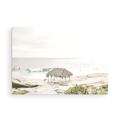Fine art of WindanSea Beach, La Jolla California horizontal landscape print by Southern California photographer Kim Dybczak of Salt Creek Prints, featuring a bright, slightly desaturated image shows Windansea Beach in La Jolla, California. A thatched beach hut stands on the sandy shore in the left foreground. Further down the beach, a surfer carrying a teal surfboard walks towards the ocean where several other surfers are visible in the water. Photograph in minimalist unframed mock up
