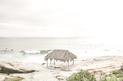 Fine art of WindanSea Beach, La Jolla California horizontal landscape print by Southern California photographer Kim Dybczak of Salt Creek Prints, featuring a bright, slightly desaturated image shows Windansea Beach in La Jolla, California. A thatched beach hut stands on the sandy shore in the left foreground. Further down the beach, a surfer carrying a teal surfboard walks towards the ocean where several other surfers are visible in the water. 