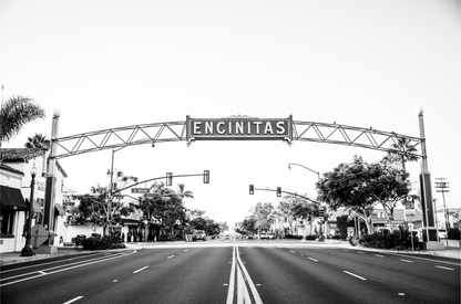 A black and white photograph of a street sign that reads 'Encinitas' with a background of a city street. 