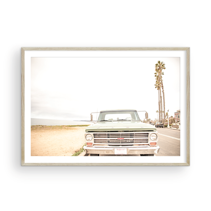 Nostalgic Southern California beach art featuring a classic light green Ford F-Series pickup truck parked by the San Diego coast. This horizontal surf print captures the vintage vehicle with a backdrop of iconic tall palm trees, a hazy beach, and the ocean, embodying surf vibes and perfect for coastal farmhouse decor. Photograph in minimalist framed mock up.